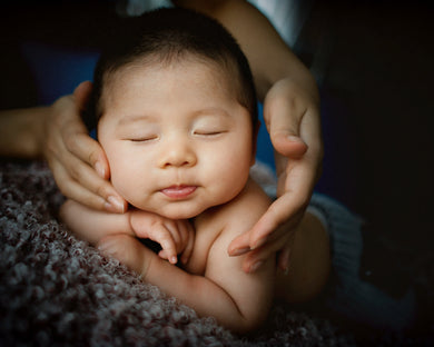 A close-up image of a baby resting on a textured surface while being held by an adult hand, which is touching the baby's ear.